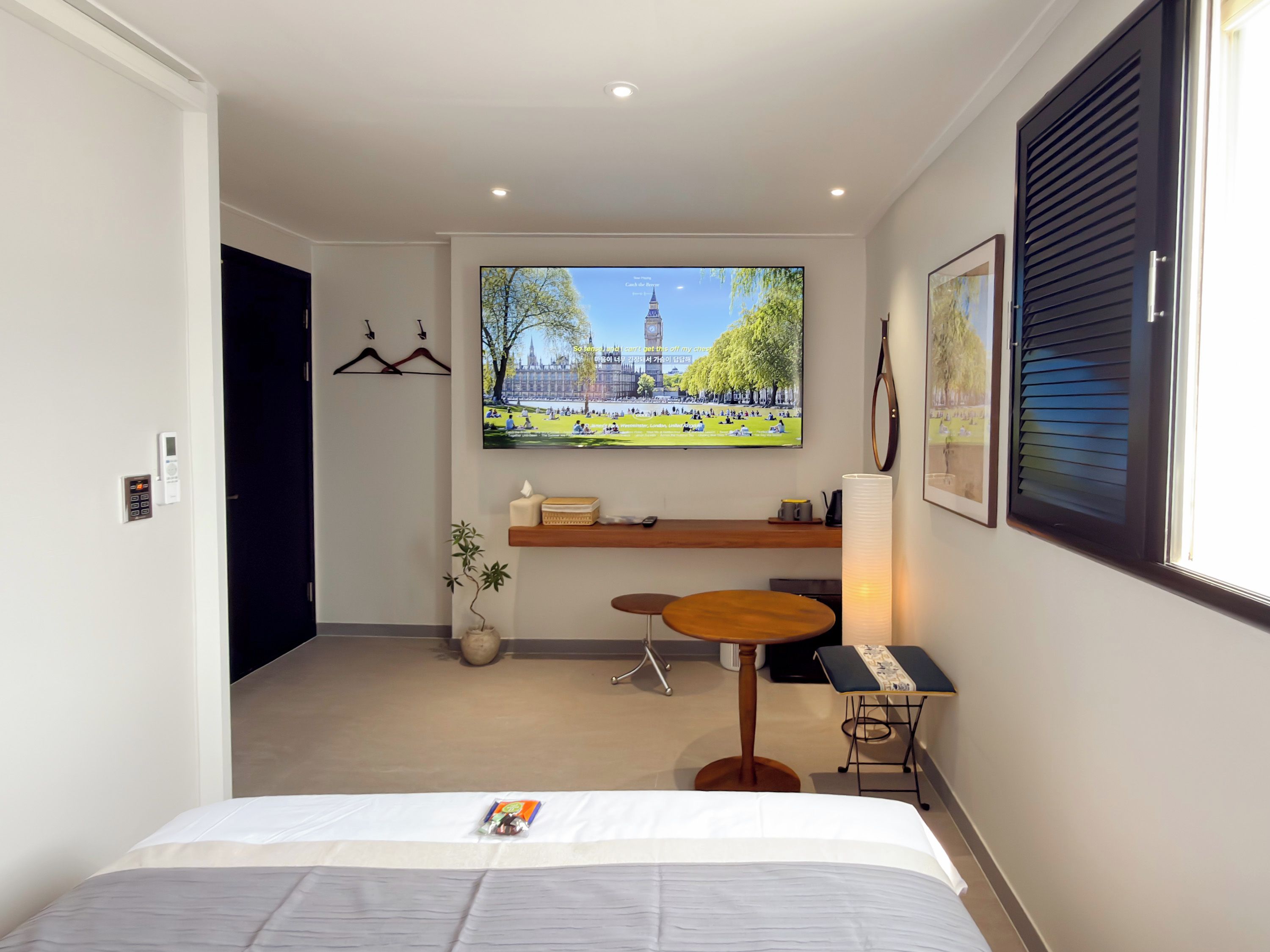 A hotel room with a bed, a TV displaying a scenic view, a small table, and a shelf with items.