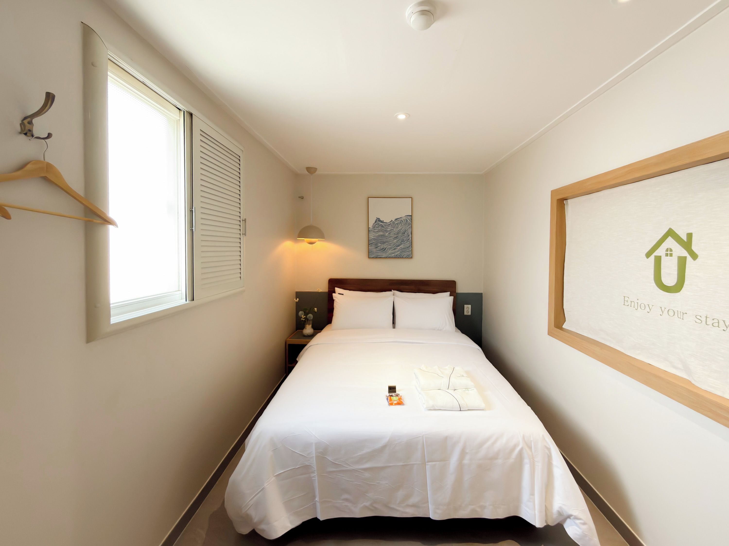 Habitación de hotel con cama, mesita de noche, perchero, un cuadro en la pared y un cartel enmarcado con un icono de casa.