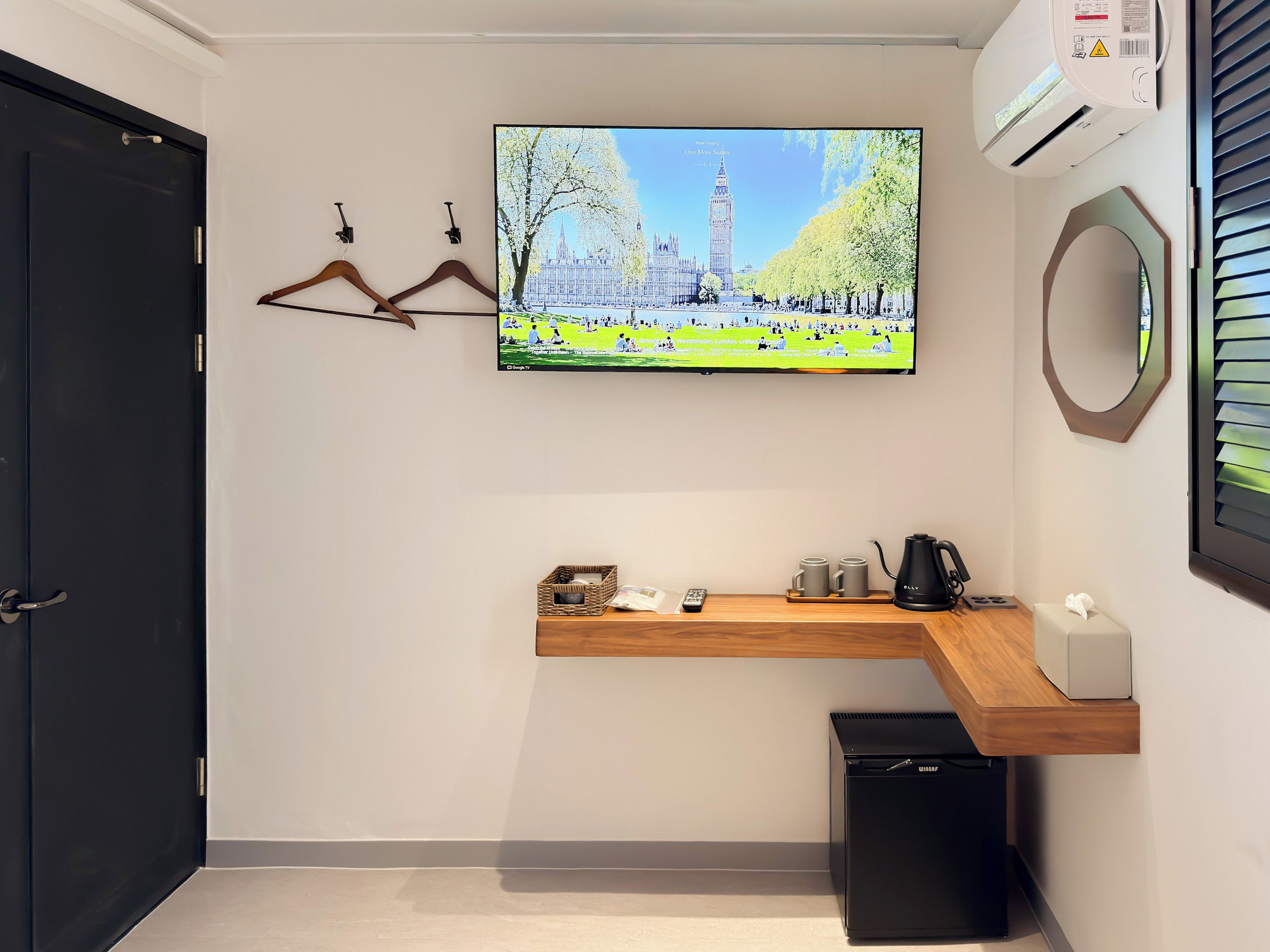 A hotel room with a flat-screen TV mounted on the wall, a wooden shelf with a kettle and mugs, a black trash bin, and a black door.