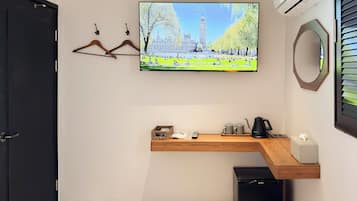 A hotel room with a flat-screen TV mounted on the wall, a wooden shelf with a kettle and mugs, a black trash bin, and a black door.