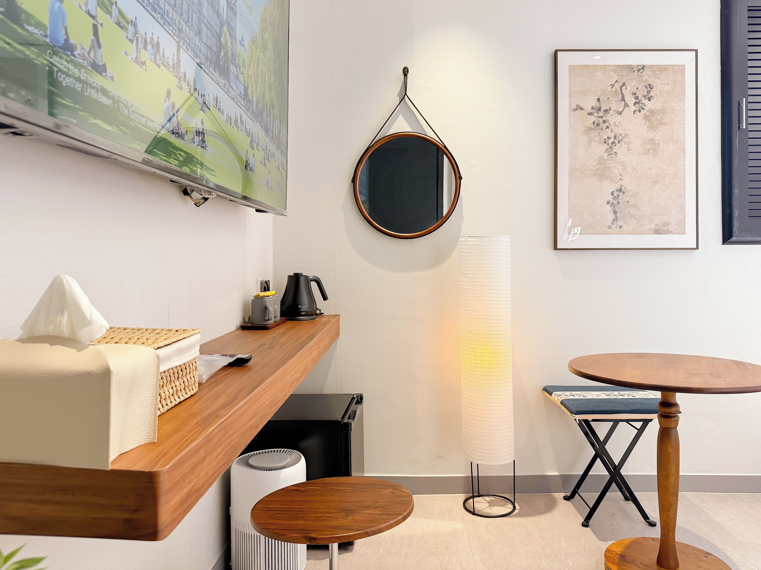 A modern hotel room with a wooden desk, a round mirror, a standing lamp, a small round table, and a wall-mounted TV.