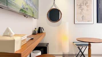A modern hotel room with a wooden desk, a round mirror, a standing lamp, a small round table, and a wall-mounted TV.