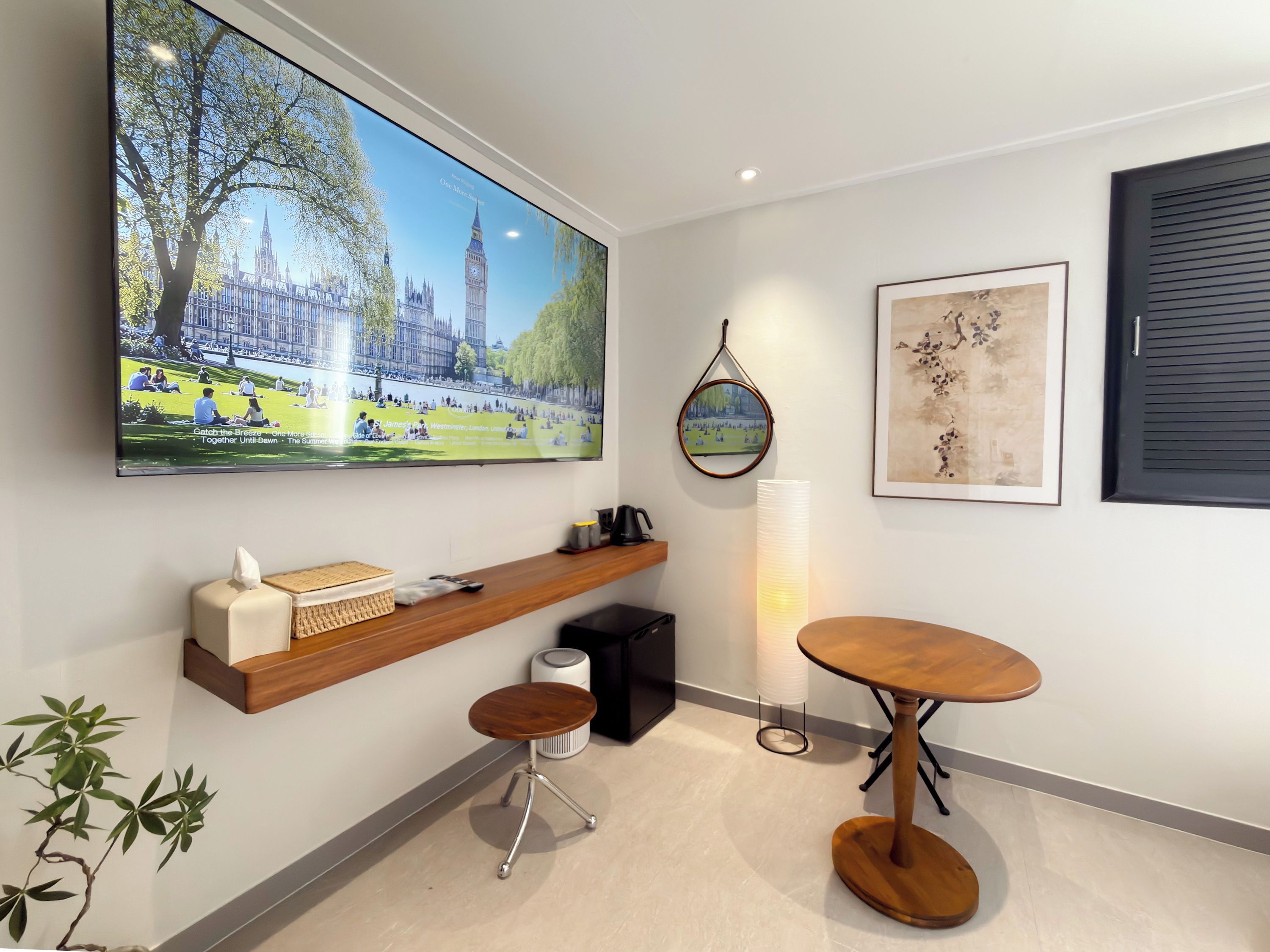 A modern room with a large TV displaying a cityscape, a wooden shelf with a tissue box, a small table, and a framed artwork on the wall.