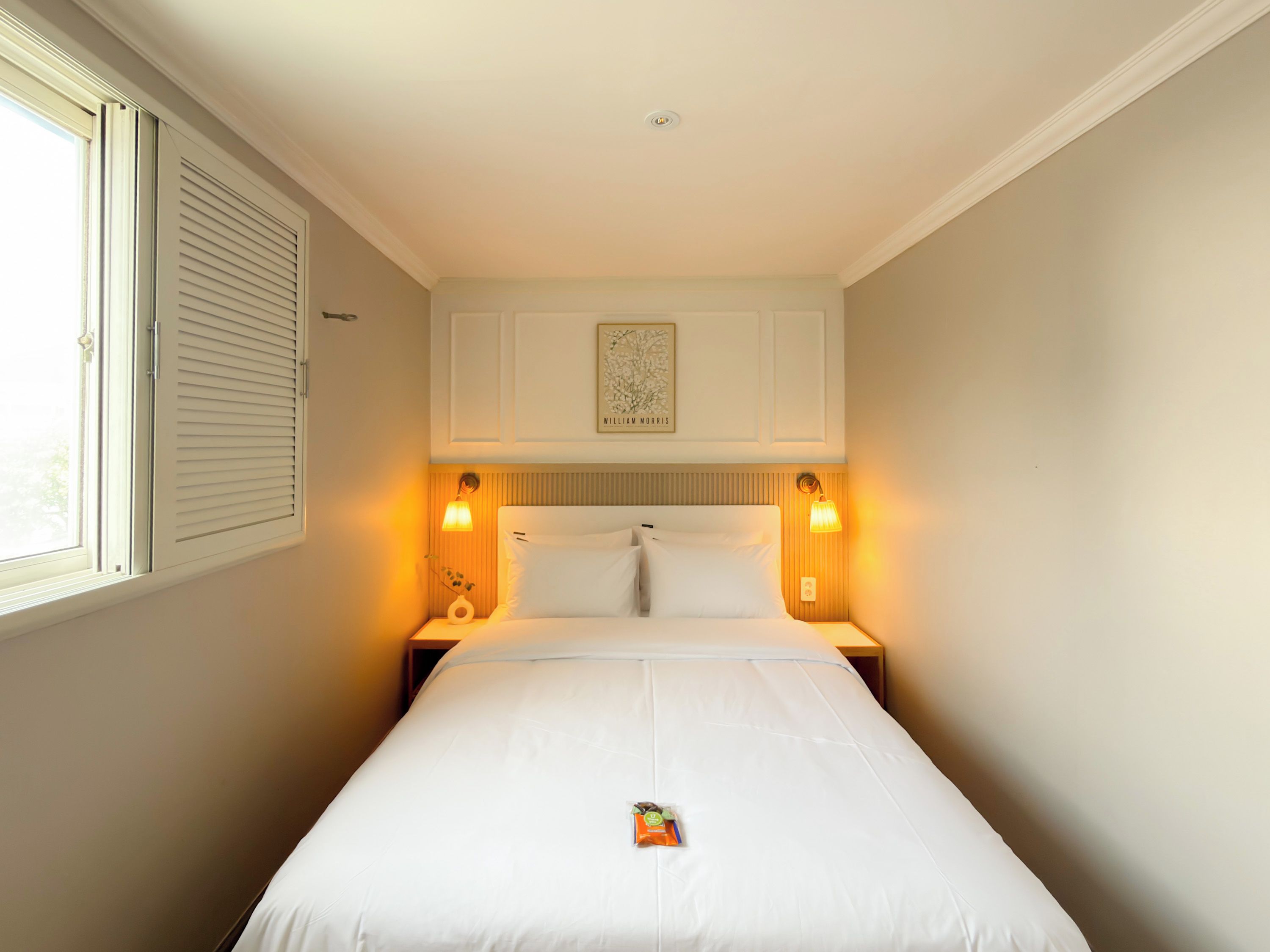 Habitación de hotel con una cama individual, dos lámparas de noche, una ventana con persianas y un cuadro en la pared.