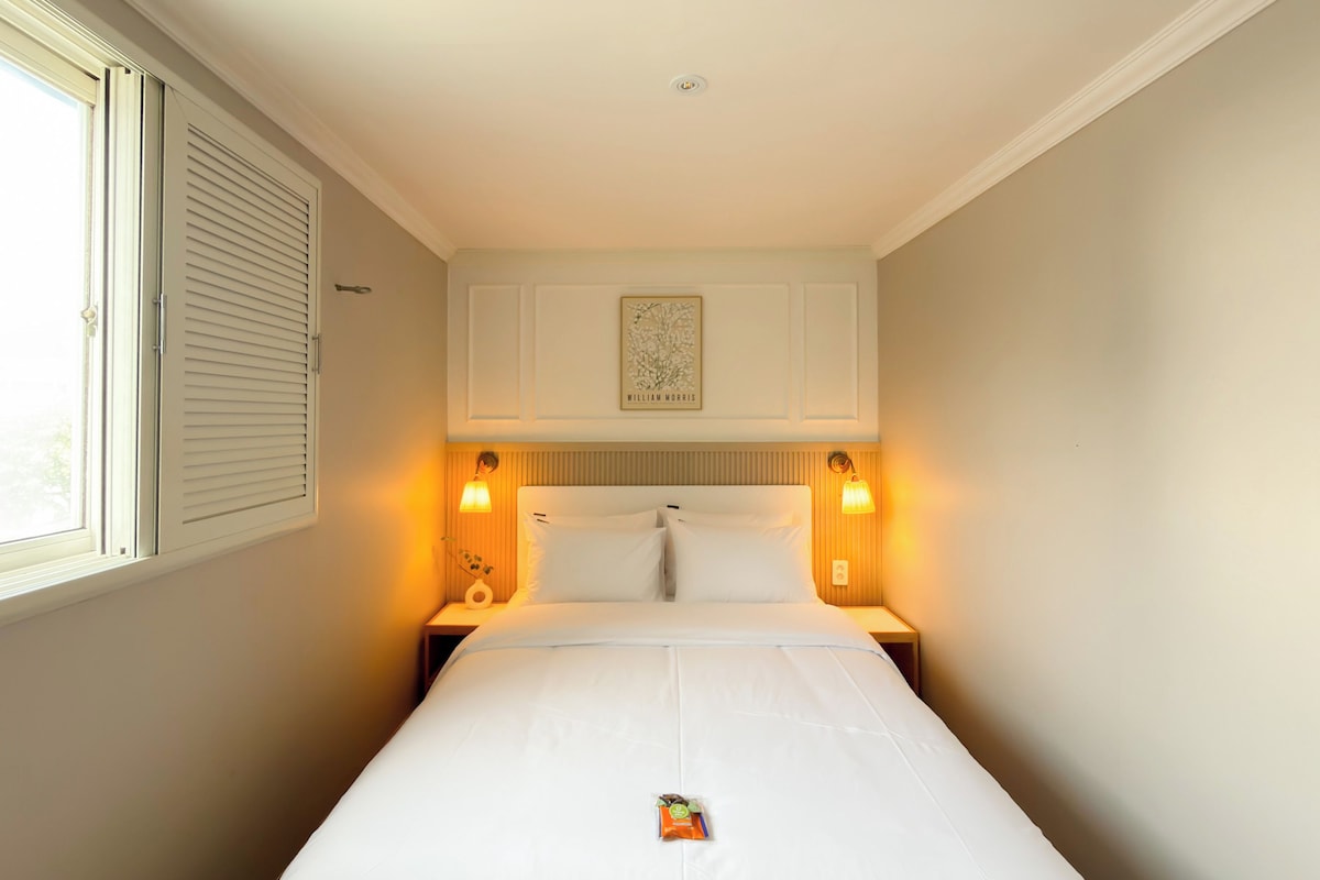 A hotel room with a single bed, two bedside lamps, a window with blinds, and a framed picture on the wall.