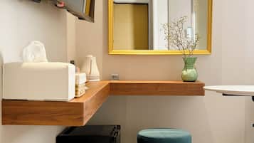 A hotel bathroom with a wooden vanity, a sink, a mirror with a gold frame, a vase with flowers, a trash bin, and a black suitcase.