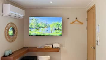 A hotel room with a flat-screen TV mounted on the wall, a wooden desk, a white chair, a small black speaker, and a door with a handle.