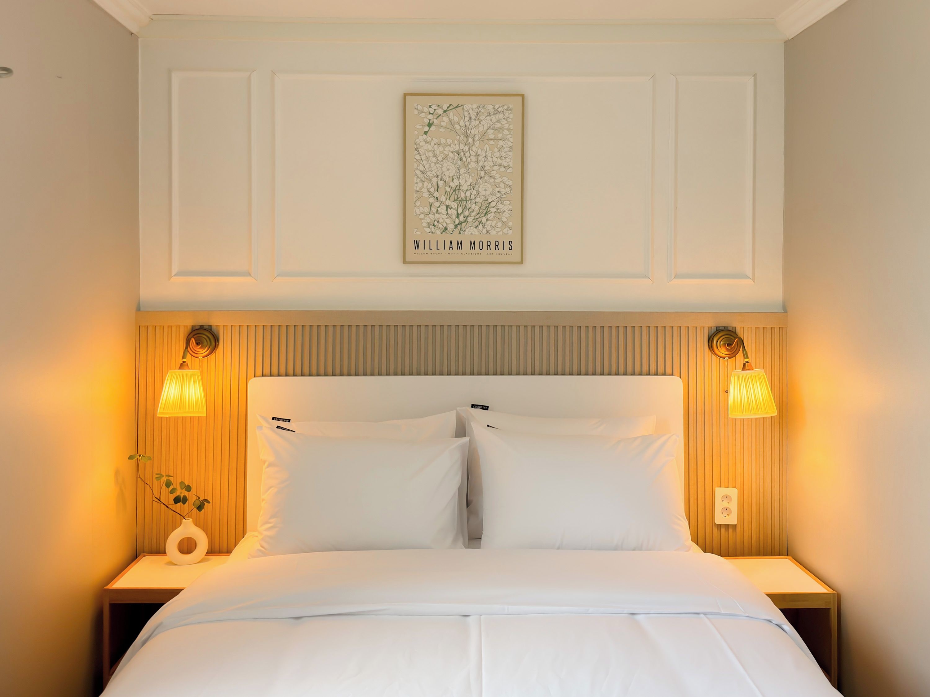 A neatly made bed with white linens, flanked by two bedside tables with lamps, and a framed artwork above the bed.
