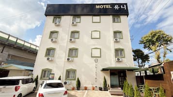 Un hôtel à plusieurs étages, avec une enseigne indiquant « Motel U ». Chaque étage est équipé de climatiseurs, l’entrée est couverte et il y a un espace de stationnement où sont garées deux voitures.