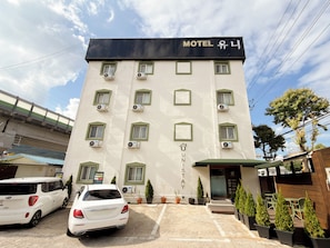 A multi-story hotel with a sign reading 'Motel U'. There are air conditioning units on each floor, a covered entrance, and a parking area with two cars. - Unistay (Dongducheon)
