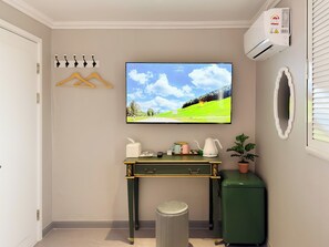 A room with a television mounted on the wall displaying a landscape, a small table with a kettle and a cup, a trash bin, and a green appliance.