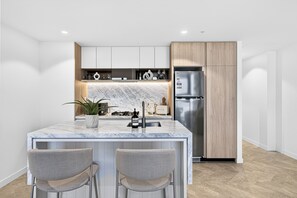 A modern kitchen with a marble countertop, stainless steel appliances, and a built-in island with bar stools. - Million Dollar Views - Luxe 62nd Floor Apartment (Brisbane City)
