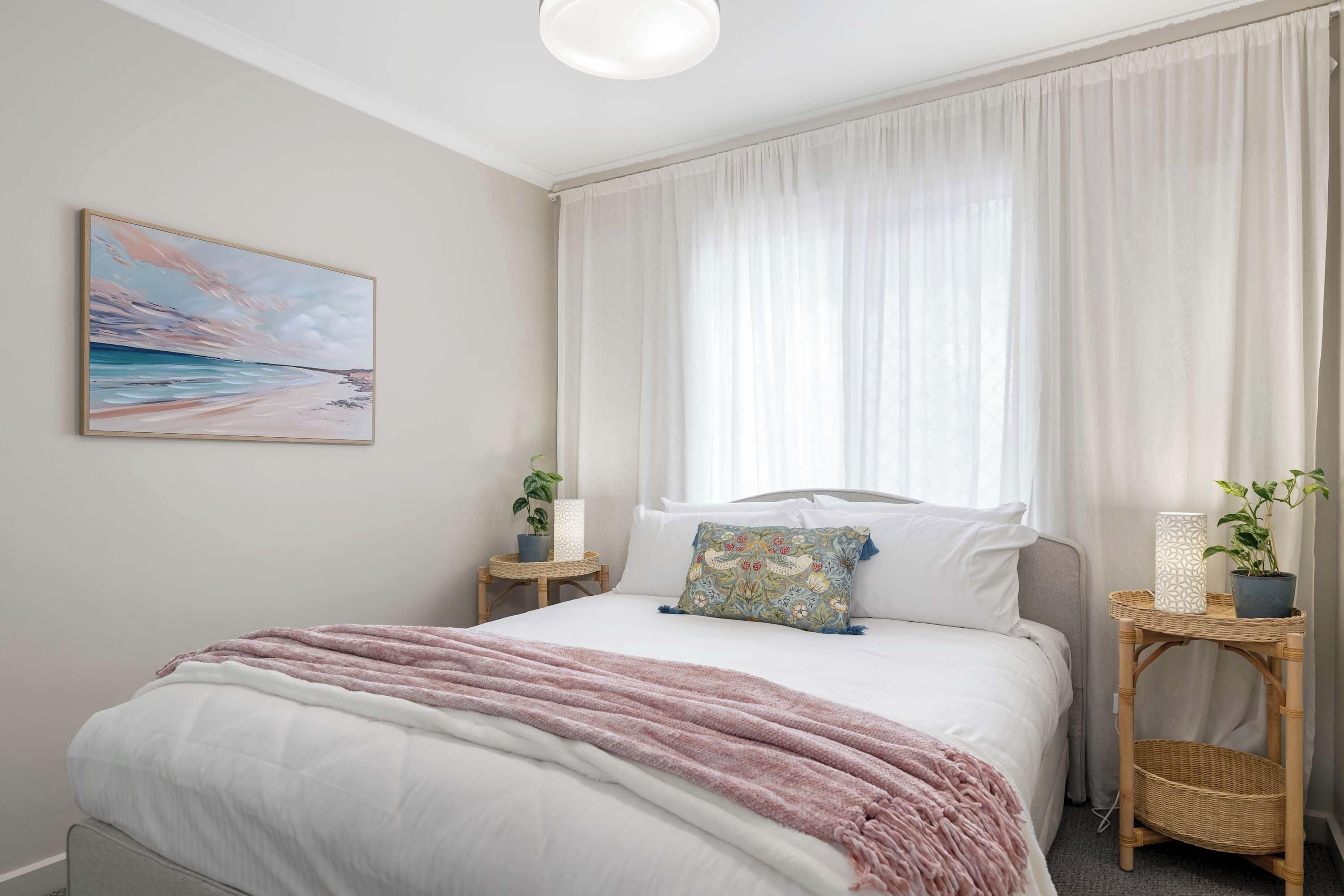 A bedroom with a bed, a nightstand, a lamp, a potted plant, and a painting on the wall.