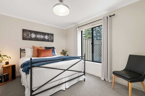 A bedroom with a bed, a chair, a window with curtains, a bedside table, and a wall-mounted artwork. - Broadleaf by the Beach | Glenelg South (Glenelg South)