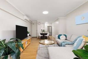 A modern living room with a flat-screen TV, a sofa, a coffee table, and a dining area with chairs. - Broadleaf by the Beach | Glenelg South (Glenelg South)