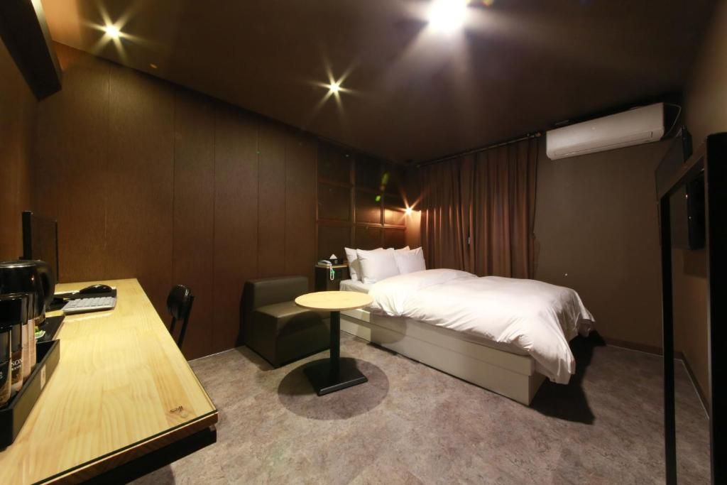 A hotel room with a bed, a desk with a chair, a small table, and a wall-mounted air conditioner.