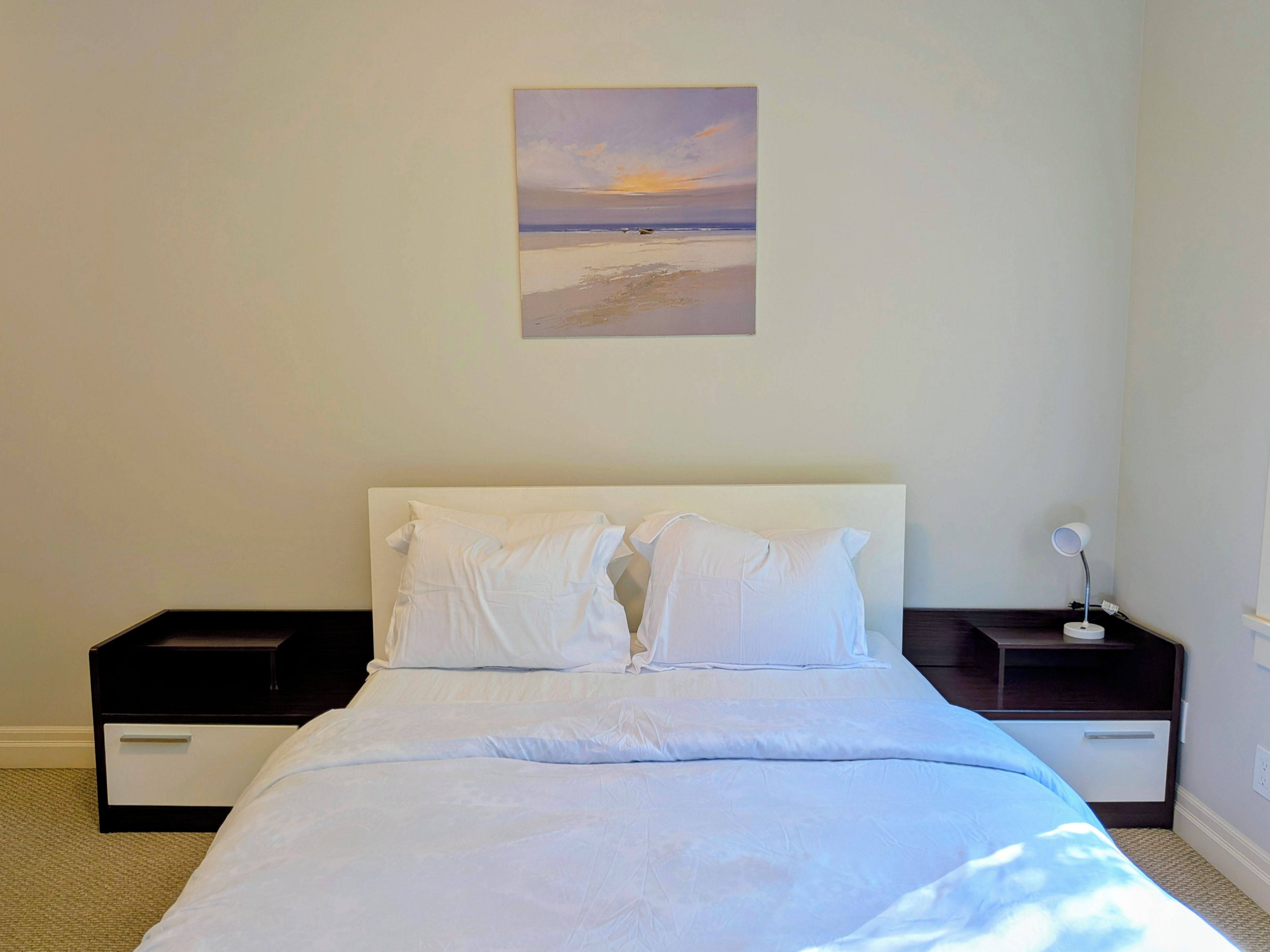 Una camera da letto con un letto, biancheria bianca, un comodino, una lampada e un quadro raffigurante una spiaggia.