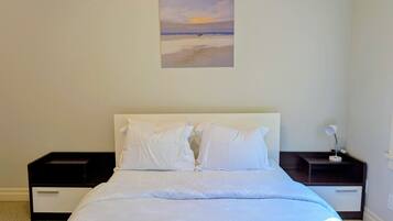 Una camera da letto con un letto, biancheria bianca, un comodino, una lampada e un quadro raffigurante una spiaggia.