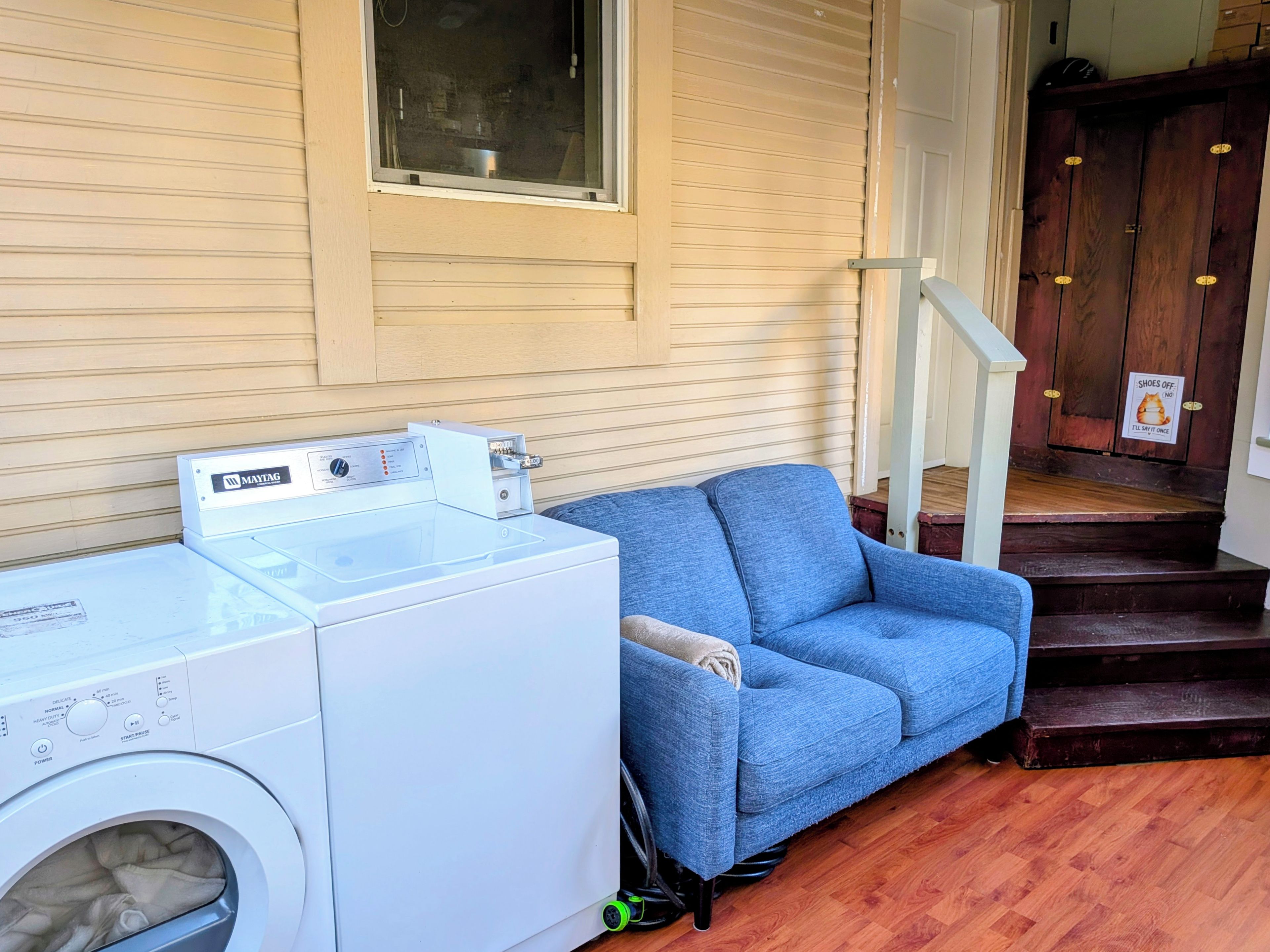 A laundry area with a front-loading washer and dryer, a blue sofa, and a staircase leading to a wooden door with a 'Shoes Off' sign.