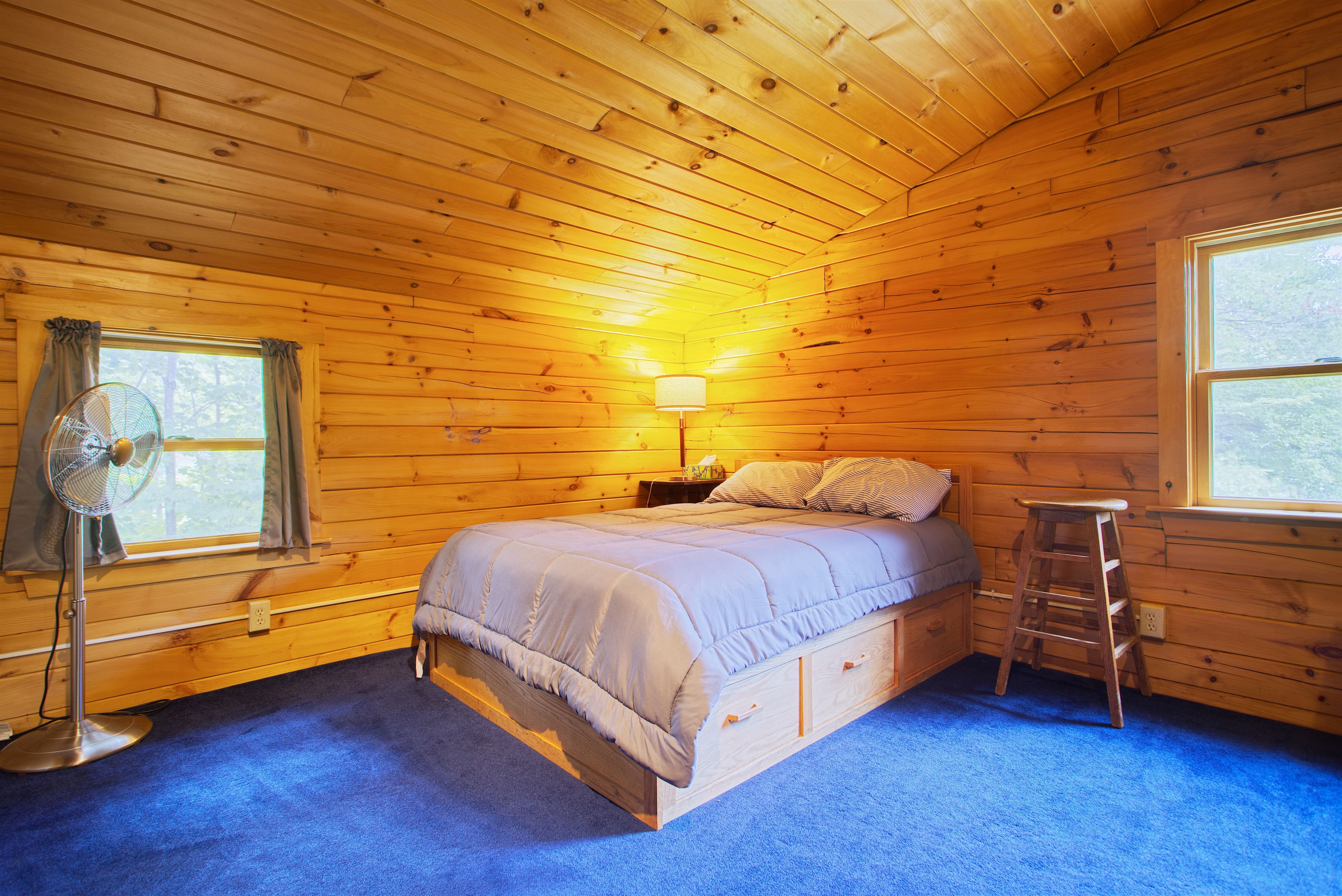 A wooden cabin bedroom with a bed, a standing fan, a nightstand, and a window.