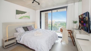Un dormitorio moderno con una cama grande, un escritorio con una computadora portátil y vistas a un paisaje urbano desde un balcón.