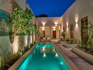 Uma piscina com superfície de água azul cristalina, rodeada por um jardim ajardinado com diversas plantas e uma área de estar com espreguiçadeiras.