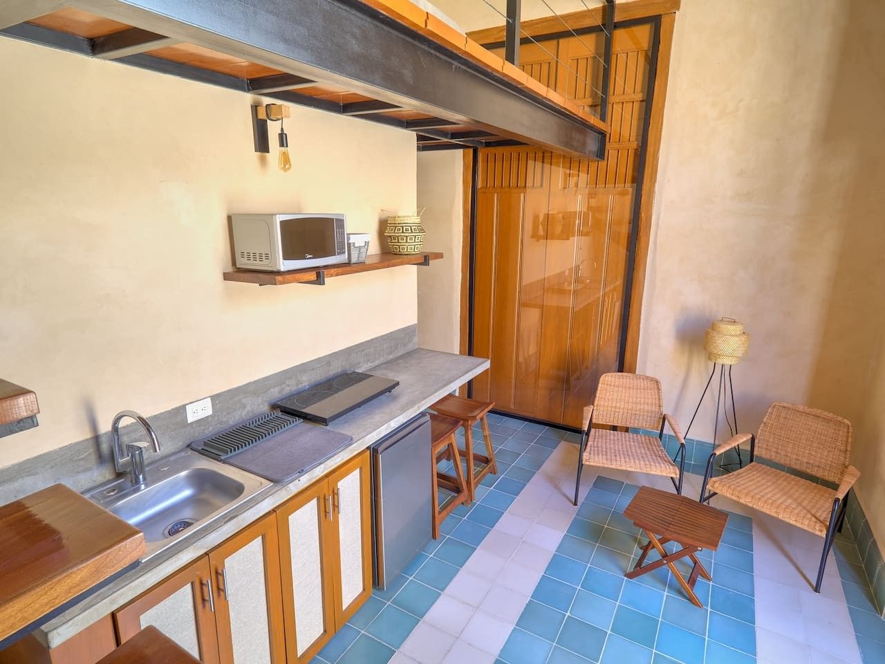 A compact kitchen with a sink, microwave, and wooden cabinets. There are two wicker chairs and a small table.