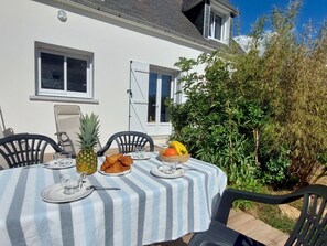 Ein Essbereich im Freien mit einem Tisch für zwei Personen, ausgestattet mit einer Tischdecke in Streifenoptik, Tellern, Besteck, einer Ananas, Croissants und einer Obstschale.