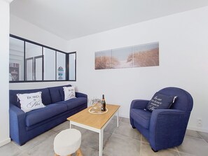 Ein modernes Wohnzimmer mit einem blauen Sofa, einem passenden Sessel, einem kleinen Holztisch mit Gläsern und einer Flasche sowie einem gerahmten Bild an der Wand.