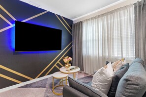 Un salon moderne avec un téléviseur à écran plat fixé au mur, un canapé avec des coussins, une petite table ronde ornée d’un vase de fleurs et une fenêtre avec des rideaux.