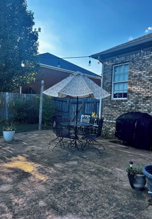 A backyard with a patio table and chairs, a patterned umbrella, a brick building, and a wooden fence.