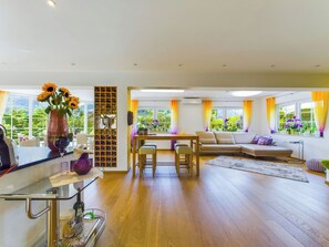 A modern living room with a wooden floor, a sofa, a dining table with chairs, and a kitchen area with a wine rack and a vase of flowers.