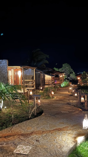 A pathway leading to a rustic building with a stone facade, wooden deck, and ambient lighting.