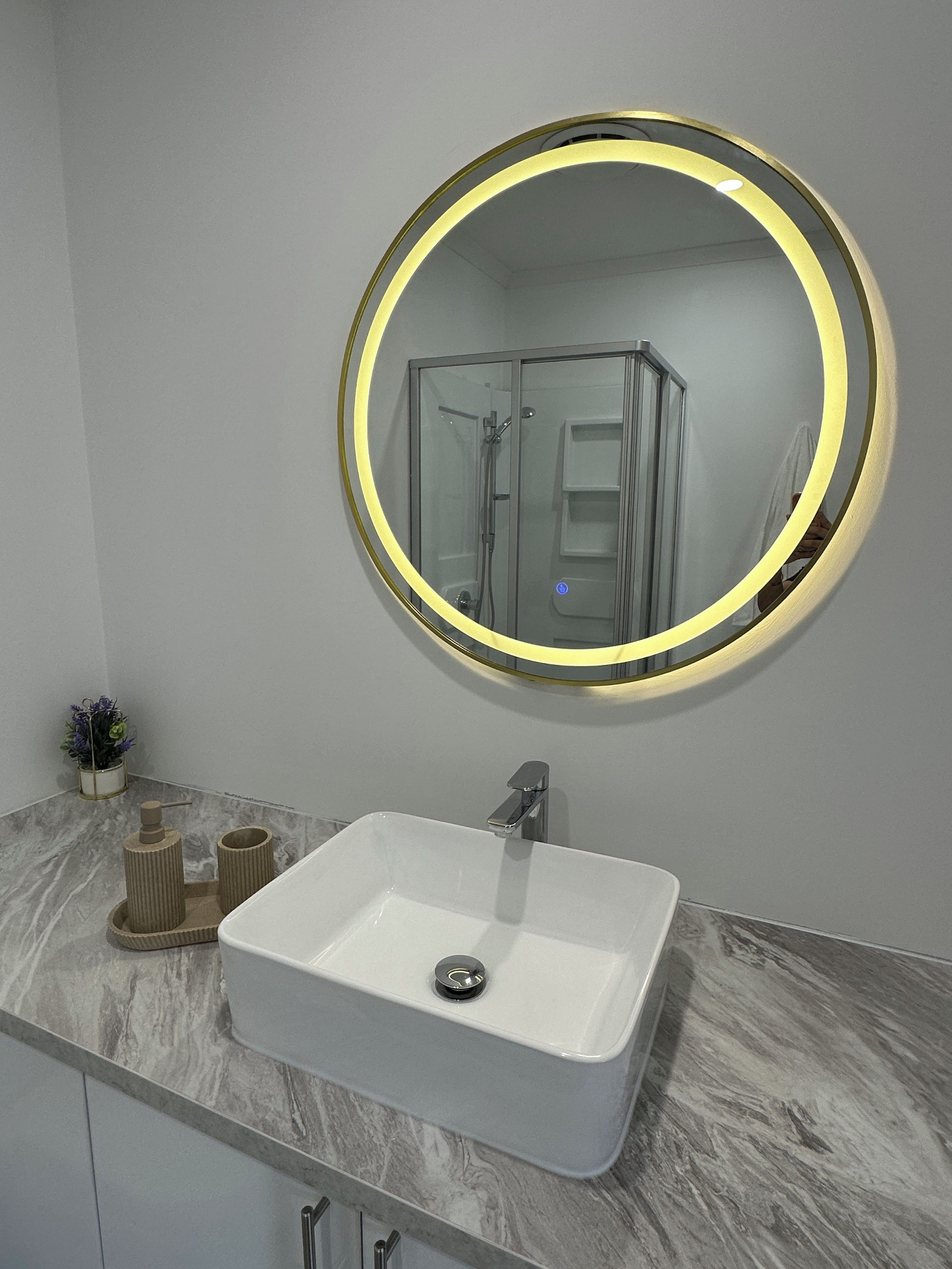 A bathroom with a white sink, a round mirror with a light, and a glass shower enclosure.