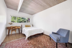 Ein Schlafzimmer mit einem Bett, einem Sessel, einem kleinen Tisch, einem Fenster mit Blick auf Grünflächen und einem gemusterten Teppich.