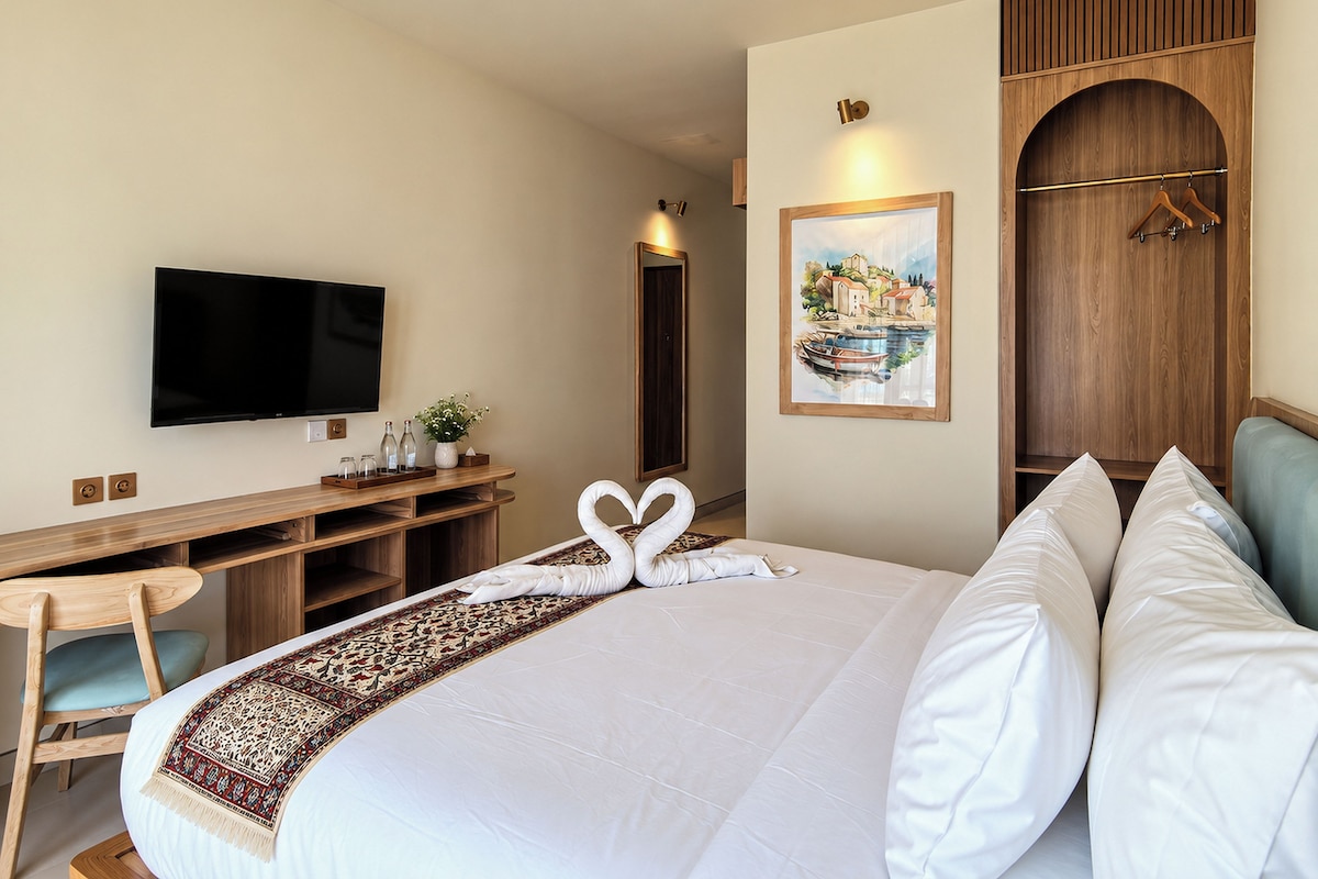 A hotel room with a large bed, a flat-screen TV, a wooden desk with a plant, and a painting on the wall.
