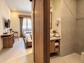 A hotel room with a wooden desk, a bed, a bathroom with a sink and mirror, and a shower area. - Laurel Davien Hotel & Resort (West Sumbawa)