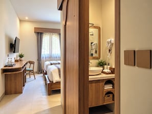 A hotel room with a wooden desk, a bed, a bathroom with a sink and mirror, and a TV mounted on the wall. - Laurel Davien Hotel & Resort (West Sumbawa)