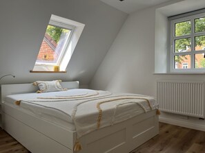 A bedroom with a sloped ceiling, a bed with a white mattress and decorative pillows, a window with a view of a brick building, and a radiator. - ------------------------urban Gem-------------------------------- (Fehmarn)