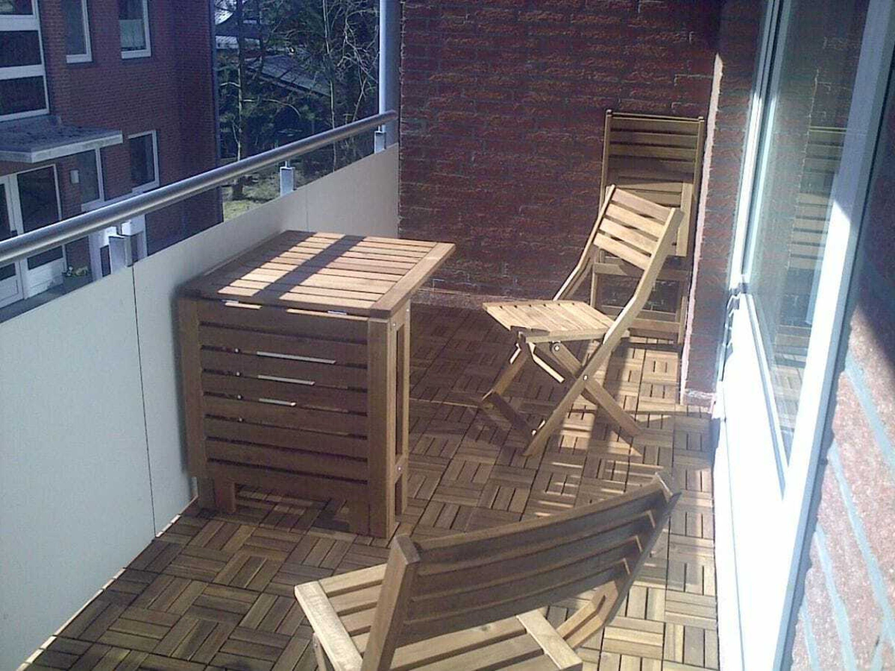 A balcony with a wooden table and chairs, a small wooden cabinet, and a view of a building and trees through a glass door.