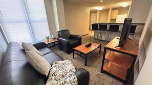A compact living space with a sofa, a coffee table, a black leather chair, and a kitchenette area with a refrigerator and bar stools.
