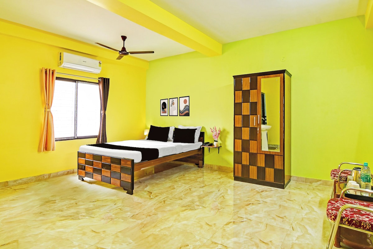 A hotel room with a bed, a ceiling fan, a window with curtains, a wall-mounted air conditioner, a bedside table with a lamp, and a mirror with a wooden frame.