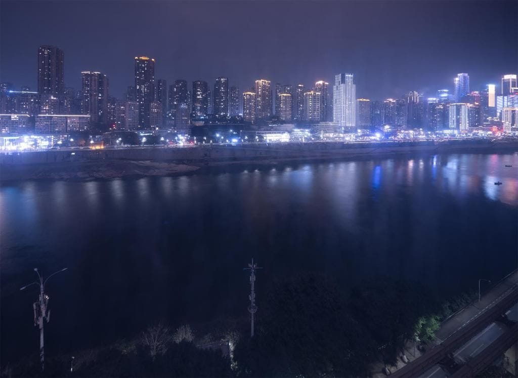 Paisatge urbà nocturn amb un riu que reflecteix llums, edificis alts i un pont.