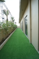 Un pasillo con piso verde artificial, una estructura metálica a la izquierda y un edificio con amplias ventanas a la derecha.