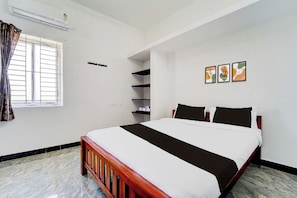 Quarto de hotel com cama, janela com cortinas, prateleira na parede e três quadros emoldurados.