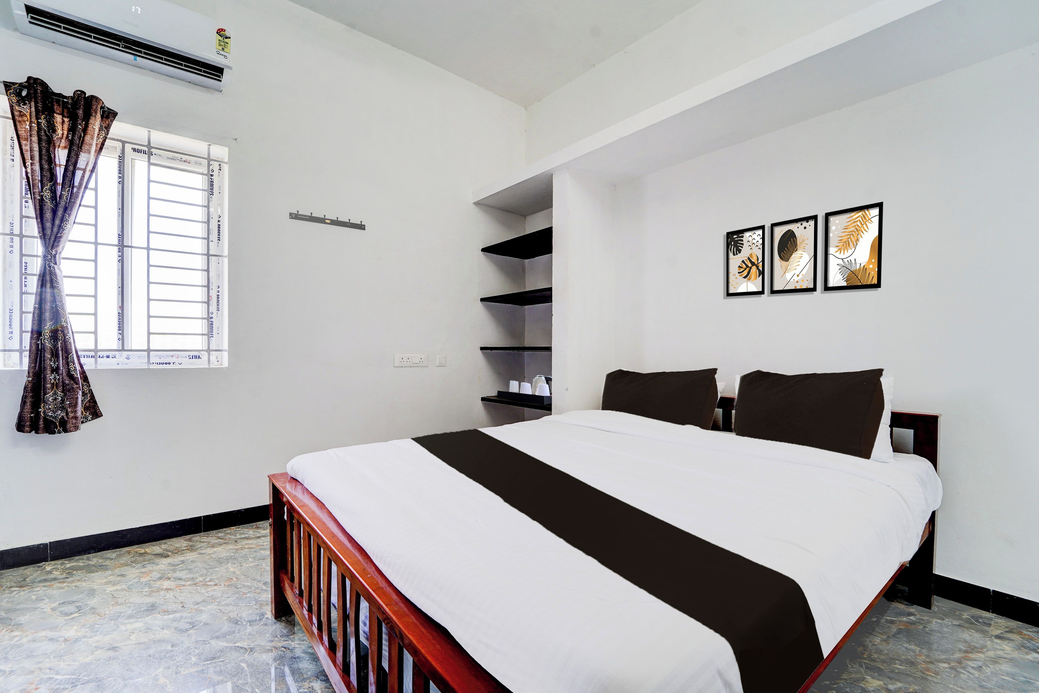 A hotel room with a bed, a window with curtains, a wall-mounted shelf, and three framed pictures.
