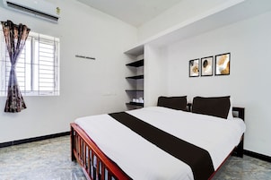 Una habitación de hotel con cama, ventana con cortinas, estante montado en la pared y tres cuadros enmarcados.