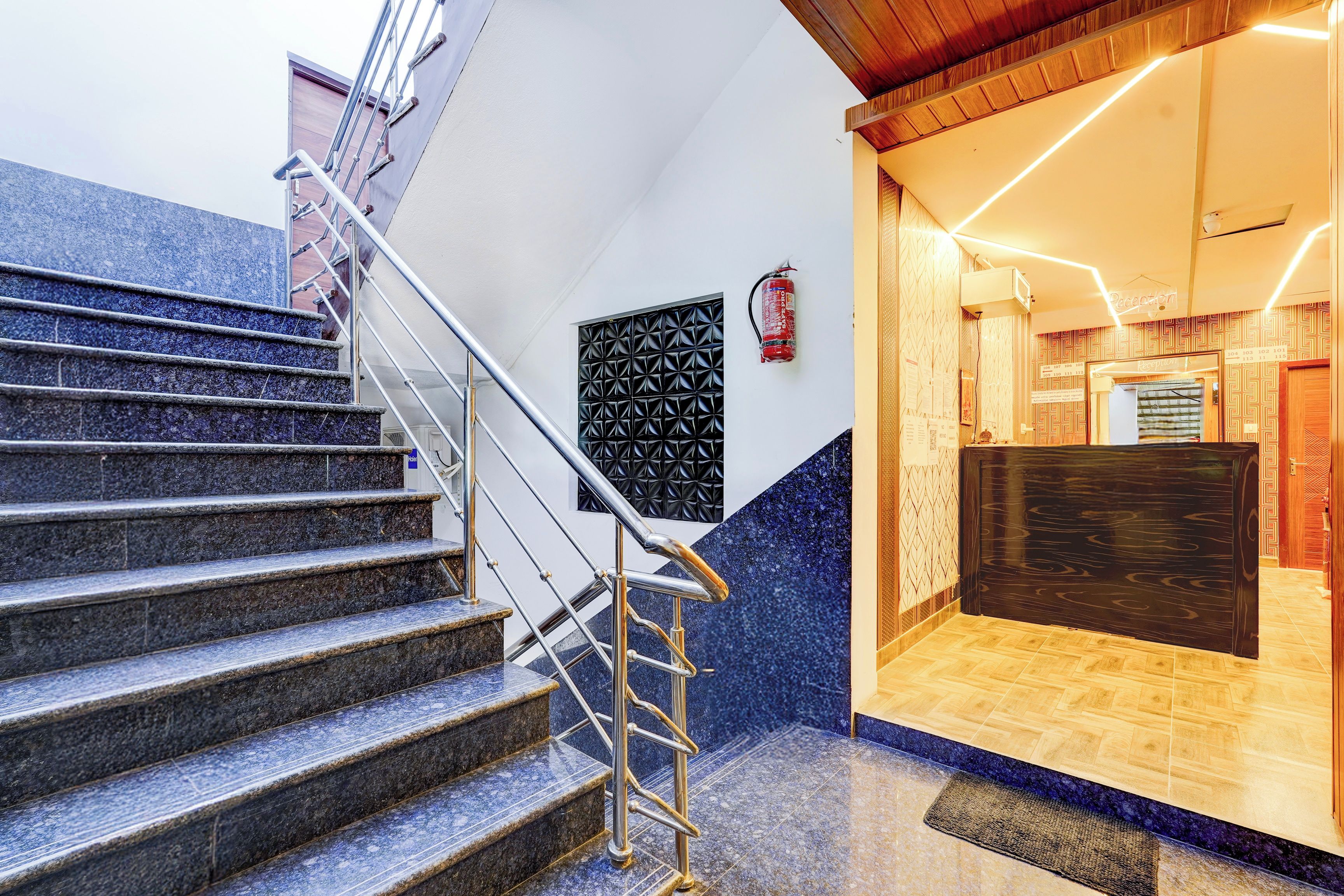 Ein Hotel-Rezeptionsbereich mit einer Rezeption, einem an der Wand befestigten Feuerlöscher und einer Treppe mit Metallgeländern, die zu einer oberen Etage führt.