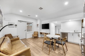 Una sala de estar moderna con un sofá, una mesa de comedor con sillas, un televisor montado en la pared y una zona de cocina con lavavajillas.