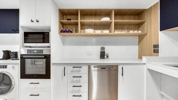 A modern kitchen with white cabinetry, a microwave, oven, dishwasher, and a wooden shelf above the sink.